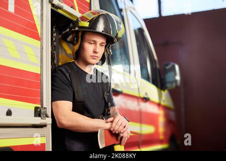 Photo d'un pompier portant un casque avec une hache contre un moteur d'incendie. Banque D'Images