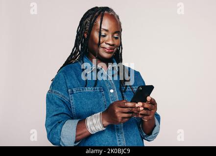 Femme d'âge moyen lisant un message texte sur un smartphone tout en se tenant debout sur un fond gris. Femme tendance avec des dreadlocks portant un denim j Banque D'Images
