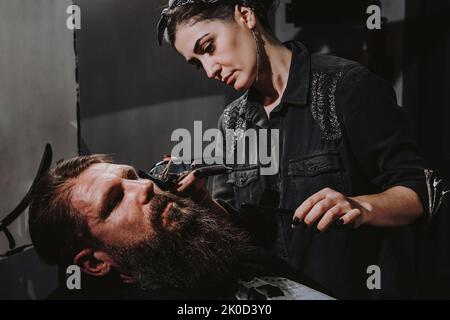 Femmes salon de coiffure professionnel servant client avec une grosse barbe épaisse par tondeuse, beau brutal jeune homme élégant obtenir une coupe de cheveux tendance, salon de coiffure noir Banque D'Images