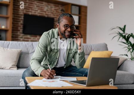 Souriant occupé mature afro-américain gars Manager dans des lunettes et décontracté, ont l'appel par téléphone, travaille sur ordinateur portable Banque D'Images