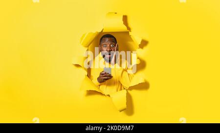 Un homme afro-américain choqué sur le plan émotionnel en utilisant un smartphone et en regardant l'appareil photo, posant dans un trou déchiré sur fond jaune Banque D'Images