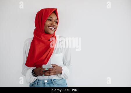 Bonne femme musulmane noire en foulard à l'aide d'un smartphone et regardant de côté l'espace libre sur fond de mur léger Banque D'Images