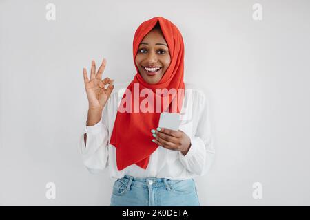 Super application. Femme musulmane noire excitée dans le hijab en utilisant un smartphone et montrant le geste ok, le fond de mur léger Banque D'Images