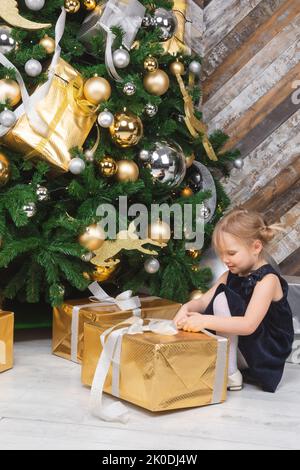 Jeune fille d'âge élémentaire portant une robe bleue assise à côté de l'arbre de noël déballage grande boîte cadeau emballée curieux d'un cadeau le lendemain de Noël Banque D'Images