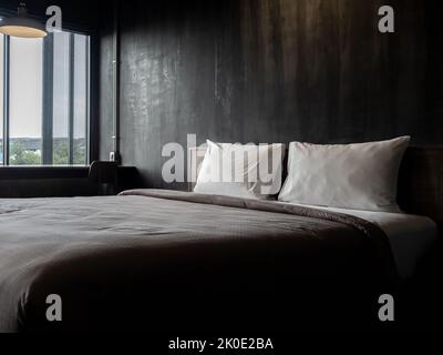 Deux oreillers blancs sur le lit vide avec couette brune sur fond de mur en béton foncé, près de la fenêtre dans le style loft de la chambre d'hôtel. Deux un doux Banque D'Images