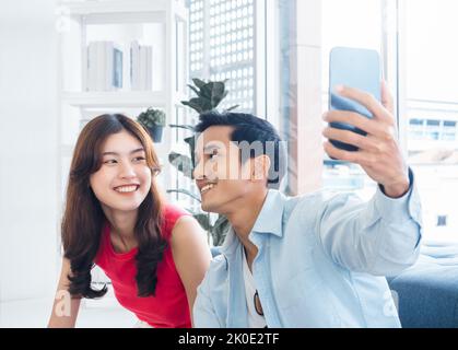Joyeux homme asiatique et jeune belle femme selfie tout en s'asseyant près de la fenêtre en verre à la salle de séjour blanche dans le bâtiment haut. Couple gai t Banque D'Images