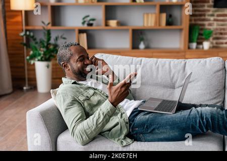 Homme afro-américain d'âge moyen souriant, pour des appels simples par téléphone, travailler avec un ordinateur portable, s'asseoir sur un canapé Banque D'Images