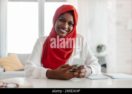 Portrait d'une femme d'affaires musulmane noire heureuse dans le hijab en utilisant le téléphone portable tout en étant assis au bureau et souriant Banque D'Images