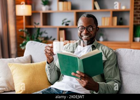 Joyeux homme afro-américain adulte dans des lunettes et livre de lecture décontracté et de profiter de la boisson préférée sur le canapé Banque D'Images