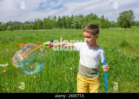 Un garçon d'âge préscolaire souffle de grosses bulles de savon dans un champ lors d'une journée ensoleillée Banque D'Images