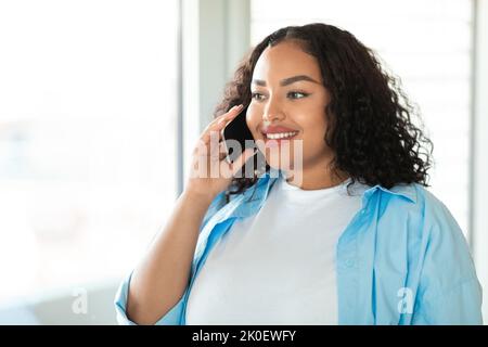 Femme noire parlant sur un téléphone portable debout près de la fenêtre à la maison Banque D'Images