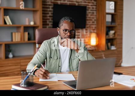 Un homme afro-américain sérieux, concentré pour les adultes, en lunettes et décontracté dans un casque sans fil fonctionne sur un ordinateur portable Banque D'Images