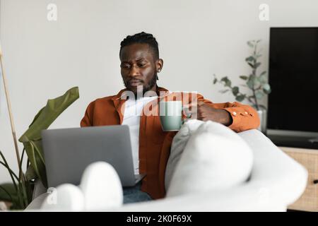 Homme afro-américain travaillant sur un ordinateur portable, tenant une tasse et prenant un café le matin, assis sur un canapé à la maison Banque D'Images