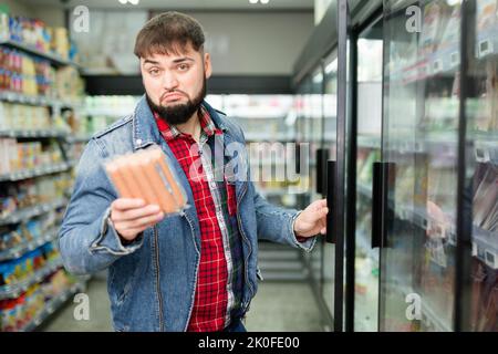 Un gars pensif à barbe qui fait des achats en magasin, en choisissant des saucisses fraîches pour le dîner Banque D'Images