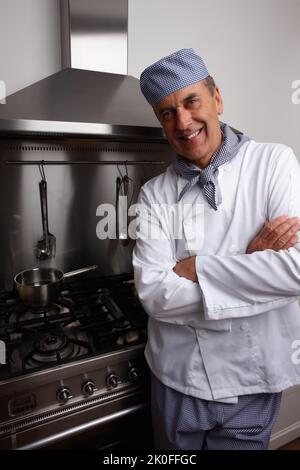 Chef cuisinier souriant avec les mains pliées debout près d'un poêle à gaz. Portrait d'un chef senior souriant avec les mains pliées debout par un poêle à gaz et une hotte d'aération. Banque D'Images