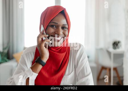 Femme noire positive dans hijab parlant sur le téléphone mobile, ayant une conversation agréable avec le client, assis au bureau à domicile Banque D'Images