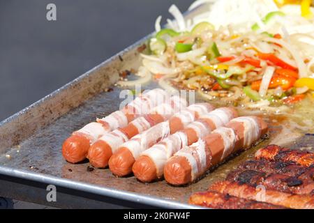 Le vendeur de rue cuisant des hot dogs enveloppés de bacon avec des oignons et des poivrons au gril à côté d'eux. Cuisine populaire pour les foires et événements de rue Banque D'Images