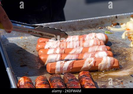 Vendeur de rue cuisant des hot dogs enveloppés de bacon, pinces tournant la nourriture. Cuisine populaire pour les foires et événements de rue Banque D'Images