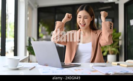 Belle et charmante jeune femme d'affaires asiatique célèbre à son bureau, regardant son écran d'ordinateur portable, ayant une nouvelle opportunité d'emploi et séquestre Banque D'Images