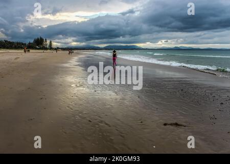 Femme avec des leggings roses et un chapeau à large bord marchant le long d'une plage humide sous un ciel orageux à Byron Bay, en Australie, avec d'autres amateurs de plage et des montagnes Banque D'Images