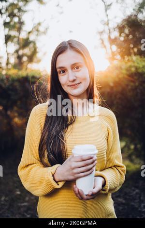 Jeune fille souriante portant un chandail jaune et tenant une tasse de café jetable au coucher du soleil dans le parc. La vibe d'automne Banque D'Images