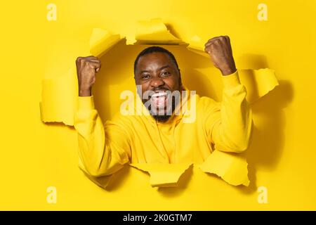 Un homme afro-américain enthousiaste qui rafle les poings, célébrant le succès et le sourire, posant dans un trou jaune déchiré Banque D'Images