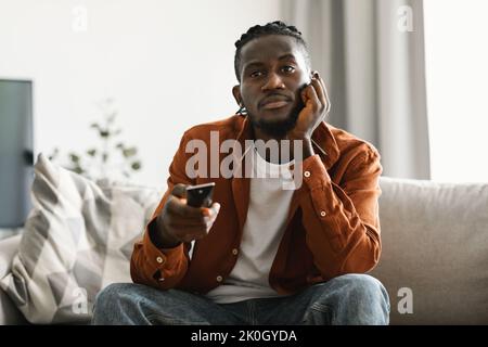 Programme de télévision ennuyeux. Ennuyé mécontent homme noir regardant la télévision et de changer de chaîne avec la télécommande, assis sur le canapé Banque D'Images