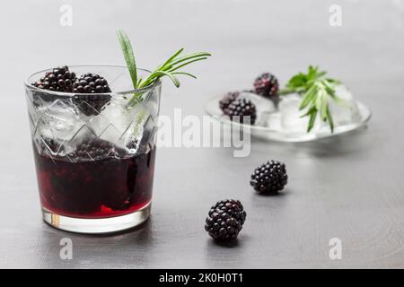 Verre traité avec de l'eau et de la glace aux mûres infusées. Glace fondue, source de romarin et baies noires sur la soucoupe. Arrière-plan gris Banque D'Images
