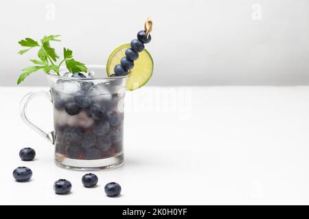 Verre traité avec de l'eau et de la glace aux myrtilles infusées. Cale de lime sur le bord du verre et bleuets sur brochette. Copier l'espace. Arrière-plan blanc. Banque D'Images