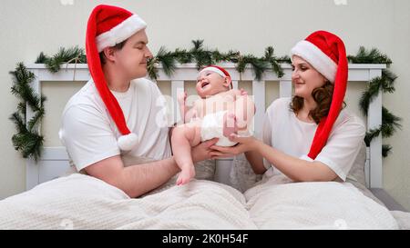 La femme donne un enfant à son mari comme cadeau sur le lit décoré pour Noël et le nouvel an, couple familial dans la chambre à coucher Banque D'Images