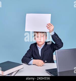 Un garçon dans un costume d'école avec une feuille de papier de A4 à un ordinateur pendant l'apprentissage à distance, un espace de copie sur un fond bleu studio Banque D'Images