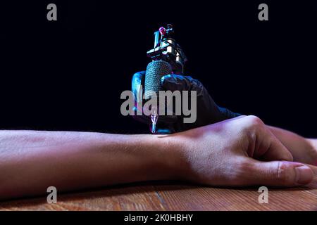 Tattaoer main de maître dans gant noir faisant l'art de tatouage sur main mâle avec machine pour tatouage sur fond sombre dans la lumière néon. Beauté, style Banque D'Images