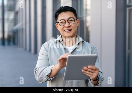 Homme d'affaires asiatique d'âge moyen heureux utilisant une tablette, regardant et souriant à la caméra, debout près d'un immeuble de bureau moderne Banque D'Images