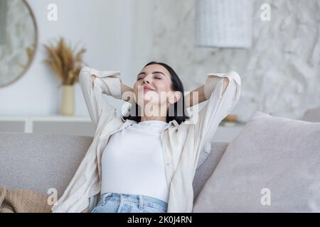 Portrait heureux jeune belle femme se reposant à la maison sur un canapé avec les yeux fermés, les bras derrière la tête. Aime le silence, la paix, les rêves, les sourires. Bonne fin de semaine. Banque D'Images