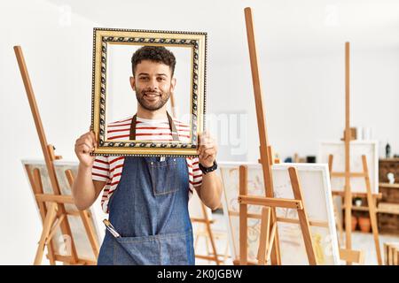 Jeune artiste arabe homme souriant heureux tenant un cadre vide au studio d'art. Banque D'Images