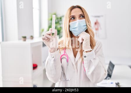 Jeune femme de race blanche, médecin, tenue d'un test covid, sérieux visage pensant à la question avec la main sur le menton, réfléchi à une idée confuse Banque D'Images