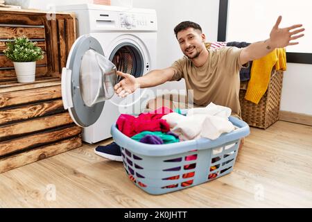 Jeune homme charmant mettant du linge sale dans le lave-linge en regardant la caméra souriant avec les bras ouverts pour le câlin. Expression gaie embrassant happ Banque D'Images