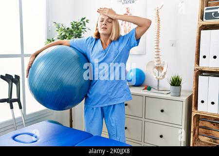 Jeune femme kinésithérapeute caucasienne tenant pilates balle stressée et frustrée avec la main sur la tête, surprise et colère visage Banque D'Images