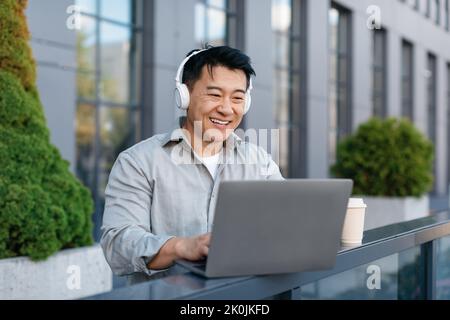 Homme d'affaires asiatique d'âge moyen heureux avec casque sans fil travaillant sur un ordinateur portable à l'extérieur, assis sur un banc Banque D'Images
