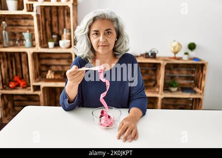 Femme d'âge moyen à cheveux gris mangeant un mètre-ruban assis sur la table à la maison. Banque D'Images