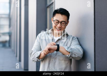 Homme d'affaires asiatique joyeux portant une montre intelligente et la regardant, en vérifiant l'heure, en utilisant des gadgets modernes à l'extérieur Banque D'Images