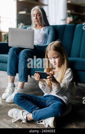 Petite-fille européenne occupée joue sur un smartphone, grand-mère âgée travaille sur un ordinateur portable sur un canapé Banque D'Images