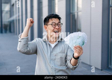 Bonhomme coréen enjogé tenant beaucoup d'argent et faisant la gestuelle OUI, homme asiatique avec ventilateur d'euros, debout à l'extérieur Banque D'Images