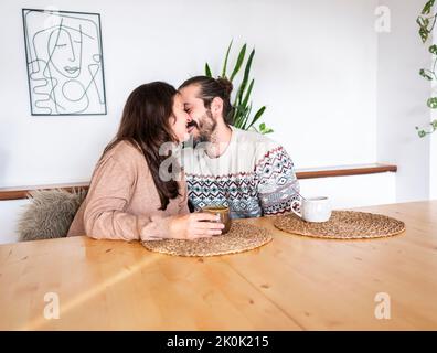 Couple aimant dans des vêtements décontractés s'embrassant tout en étant assis à table avec des tasses de café dans la pièce lumineuse Banque D'Images