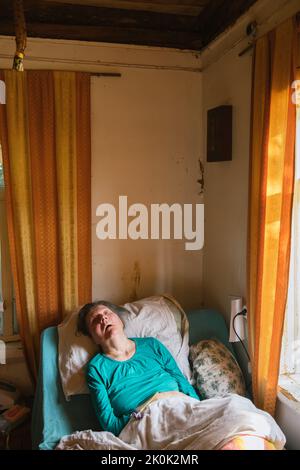 Femme âgée paralysée, couchée sous une couverture sur un lit médical près des fenêtres dans une pièce miteuse à la maison Banque D'Images