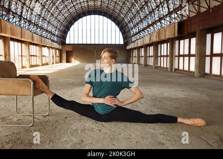 Gracieuse danseuse de ballet jeune dans le léopard noir en effectuant des fractionnements longitudinaux mettant un pied sur la chaise dans le hall vide avec de grandes fenêtres et g Banque D'Images