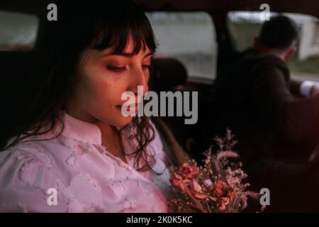 Vue de la charmante mariée souriante en robe blanche et avec bouquet floral assis en voiture avec une lumière rouge fluo contre un marié flou le jour du mariage Banque D'Images