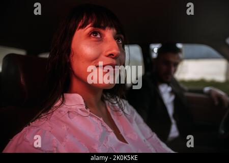 Vue de la charmante mariée souriante en robe blanche et avec bouquet floral assis en voiture avec une lumière rouge fluo contre un marié flou le jour du mariage Banque D'Images