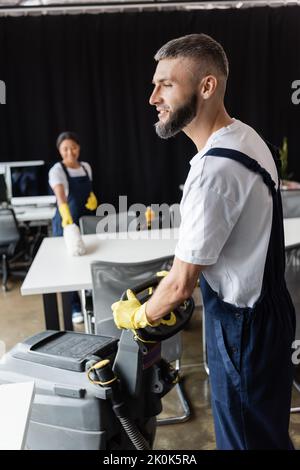 homme barbu avec machine à récurer le sol près de la race mixte femme essuyant le bureau sur un arrière-plan flou, image de stock Banque D'Images