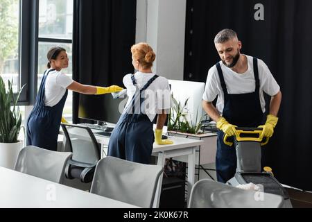 jeune femme biraciale essuyant l'écran d'ordinateur près de collègues nettoyage bureau, image de stock Banque D'Images